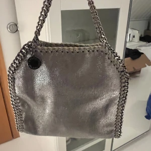 Silver axelväska från Stella McCartney - Snygg silverfärgad axelväska från Stella McCartney med kedjedetaljer längs kanterna. Väskan har en metallisk look och en praktisk magnetknäppning. Den långa kedjan ligger i väskan på bilderna. Om intresse finns, kan jag skicka bilder när jag bär väskan som crossbody!