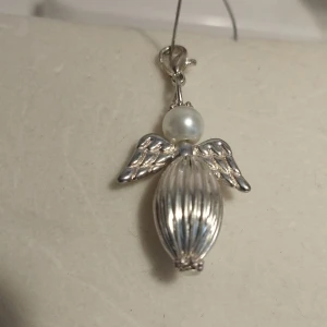Silverfärgat änglahänge med pärldetalj - Charmigt hänge i form av en lyckoängel med silverfärgade vingar och en pärla som huvud. Kroppen är avlång och räfflad, vilket ger en elegant touch. Perfekt som ett unikt smycke eller present. Cirka 4 cm.