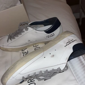 Vita sneakers från golden goose - Snygga vita sneakers med marinblå detaljer och grå snörning. Skorna har en unik design med text och koordinater tryckta på sidan. Perfekta för en stilren och modern look. 