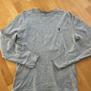 Grå långärmad tröja från Polo Ralph Lauren - Säljer en stilren grå långärmad tröja från Polo Ralph Lauren. Tröjan är tillverkad i 100% bomull och har en liten broderad logotyp på bröstet. Perfekt för en avslappnad och bekväm stil.