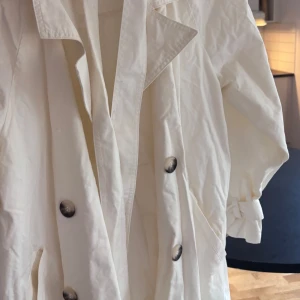 Vit trenchcoat med knappar - Snygg vit trenchcoat med knappar och klassisk krage. Jackan har långa ärmar och ett bälte i midjan för en stilren look. Perfekt för vår och höst.