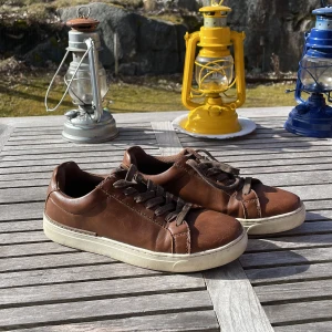 Bruna sneakers i skinn - Säljer ett par bruna sneakers i skinn med snörning. De har en vit sula och en klassisk design som passar till många olika stilar. Perfekta för en avslappnad look. Den högra skon är lite skadad men vänstra är hel!