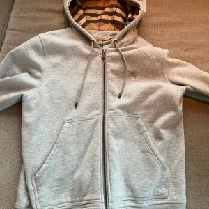 Grå hoodie från Burberry - Säljer en stilren grå hoodie från Burberry med klassiskt rutigt mönster i huvan. Tröjan har dragkedja och två fickor framtill. Perfekt för en grisch och Laidback look.