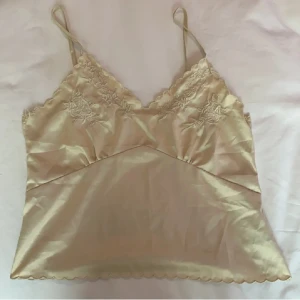 Beige sidenlinne med spetsdetaljer - Säljer ett elegant beige linne i siden med tunna axelband och vacker spetsdetalj vid halsringningen. Perfekt för en stilren och sofistikerad look. Passar bra till både jeans och kjol.