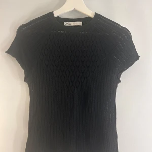 Svart topp från Zara - Snygg svart stickad topp från Zara med korta ärmar och ett elegant mönster. Perfekt för att lägga till en touch av stil till din garderob. Passar bra till både jeans och kjol.
