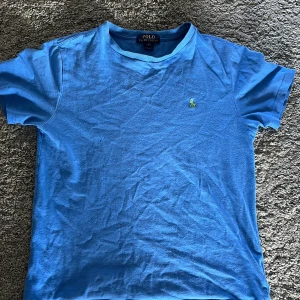 Blå t-shirt från Ralph Lauren - Säljer en klassisk blå t-shirt från Ralph Lauren med korta ärmar och en liten neongrön broderad logga på bröstet. Perfekt för en avslappnad stil. 