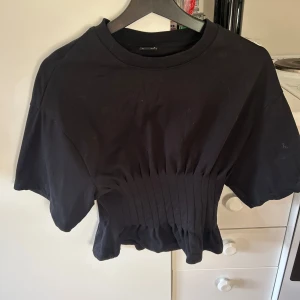 Svart kortärmad topp med ribbad midja - Snygg svart topp med korta ärmar och en unik ribbad detalj i midjan som ger en smickrande passform. Perfekt för en stilren look.