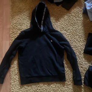 Svart hoodie och byxor från Calvin Klein Jeans - Snygg svart hoodie från Calvin Klein Jeans med logotypdetaljer på huvan. Snygga matchande byxor tillkommer också storlek 152cm.