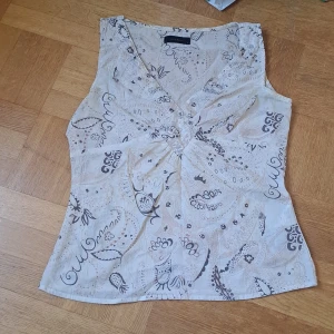 Beige mönstrad topp - Säljer en beige topp från Vero Moda med v-ringning och ärmlös design. Toppen har ett paisleyinspirerat mönster i bruna och grå toner samt små glittriga detaljer framtill. Några pärlor/paljetter sitter lite löst. Men går att fixa. 🩷🩷
