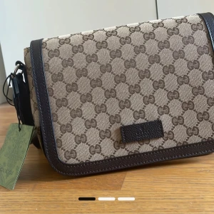 Beige axelväska från Gucci - Snygg beige axelväska från Gucci med klassiskt GG-mönster i brunt. Väskan har mörkbruna skinnkanter och justerbar axelrem. Perfekt storlek för det viktigaste och en ikonisk detalj till din outfit.