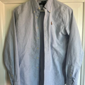 Ljusblå skjorta från Ralph Lauren - Klassisk ljusblå skjorta från Ralph Lauren med button down-krage och broderad logga på bröstet. Skjortan har lång ärm, knappar framtill och är tillverkad i bomull. Perfekt för en clean och stilren look. Skjortan i storlek S. Som ni kan se på bilden så finns det en liten fläck som knappt är synlig, tveka inte att fråga om ni vill ha fler bilder eller undrar något!
