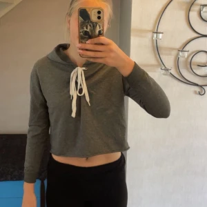 Grå croppad hoodie från Colorbox - Grå croppad hoodie från Colorbox med vit snörning framtill och huva. Tröjan har långa ärmar och är tillverkad i mjuk bomullsmix. Perfekt för en chill och avslappnad stil.