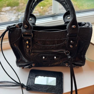 Svart  handväska i skinn - Cool svart handväska i skinn med silverfärgade nitar och dragkedja framtill. Väskan har dubbla handtag, avtagbar spegel och flera fack inuti för smidig organisering. Perfekt storlek för det viktigaste och en edgy vibe till din outfit.