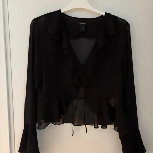 Svart volangblus från Lindex - Supersnygg svart blus från Lindex i transparent, lätt material. Blusen har långa ärmar med volangdetaljer, v-ringning och knyt framtill. Croppad modell med volang längs kanten och ärmslut. Perfekt för dig som gillar trendiga och feminina detaljer.