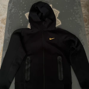 Nike NOCTA svart hoodiejacka - Svart hoodiejacka från Nike NOCTA med gul swoosh och broderade detaljer. Jackan har hel dragkedja, två fickor med dragkedja och huva. Snygga NOCTA-loggor och brodyr på ärm och rygg. Perfekt för dig som gillar streetwear och sportig stil.