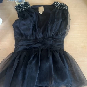 Svart blus med pärldetaljer - Supersnygg svart blus från Party Pieces med voluminöst tylltyg och markerad midja. Axelpartierna är dekorerade med svarta och silvriga pärlor för en extra glam känsla. Perfekt för dig som vill sticka ut med coola detaljer och elegant look.