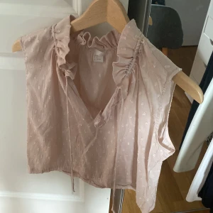 Ljusrosa blus med volang från H&M - Supersöt ljusrosa blus från H&M med volangdetaljer vid kragen och v-ringning. Blusen är ärmlös och har små broderade prickar i tyget samt knytband framtill. Perfekt för en romantisk och feminin look.
