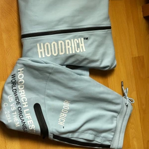 Ljusblå mjukisbyxor från Hoodrich - Snygga ljusblå mjukisbyxor från Hoodrich med tryck på benet och coola svarta detaljer vid fickorna. Byxorna har dragskedjor på sidorna  och muddar vid bensluten för en skön och avslappnad passform. Perfekta för chill eller streetwear. Koftan L, Byxorna M. 2500 orginal pris