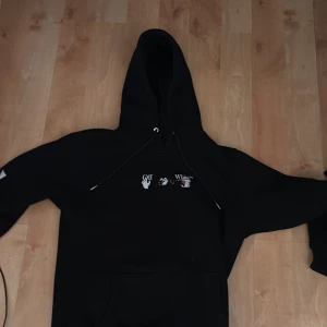 Svart Off-White hoodie med tryck - Svart hoodie från Off-White med stor huva och snörning. Framsidan har ett unikt grafiskt tryck med händer och texten 'Off White', medan ryggen pryds av ett stort konstnärligt motiv i vitt, rosa och rött. Perfekt för dig som gillar streetwear och vill sticka ut.