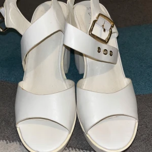 Vita sandaler med klack och spänne - Snygga vita sandaler med breda remmar och chunky klack. De har öppen tå och ett guldigt spänne som detalj. Perfekta för dig som vill ha en stilren och trendig look med lite extra höjd. Gjorda i syntetmaterial.