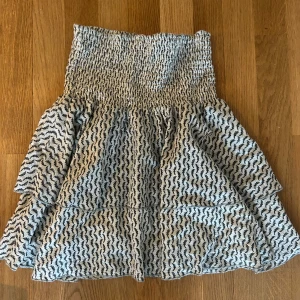 Volangkjol från Sofie Schnoor - Supersöt kortkjol från Sofie Schnoor i vitt med mörkgröna mönster och glittriga detaljer i silver. Kjolen har smockad midja och volangig nederdel som ger en luftig känsla. Perfekt för dig som gillar unika och trendiga plagg. Den är i storlek XS och tillverkad i bara bomull.  💗