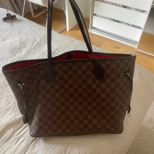 Louis Vuitton Damier Ebene shoppingväska - Säljer en klassisk Louis Vuitton Neverfull i Damier Ebene-mönster. Väskan är brunrutigt med mörkbruna skinnhandtag och detaljer. Insidan är röd med svarta ränder och har en innerficka med dragkedja. Perfekt storlek för att få plats med allt du behöver.