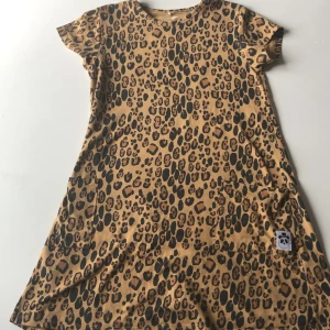 Leopardmönstrad klänning gjord av Mini Rodini - Cool kort klänning från Mini Rodini i beige med svart och brunt leopardmönster. Tillverkad i mjuk bomull, har korta ärmar och två fickor framtill. Perfekt för dig som gillar djurmönster och vill sticka ut lite extra.