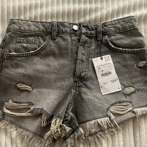 Grå jeansshorts med slitningar från Zara - Gråa midwaist jeansshorts från Zara med råa, fransiga kanter och slitna detaljer framtill och baktill. Klassisk femficksmodell med dragkedja och knapp. Perfekta för en avslappnad och trendig look i sommar.