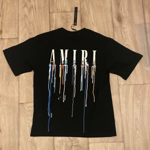 Svart AMIRI t-shirt med färgstänk - Svart t-shirt från AMIRI med stort tryck på ryggen och mindre tryck på bröstet. Trycket består av AMIRI-loggan med färgstänk i blått, vitt och orange som rinner ner. Klassisk passform och rund hals. Perfekt för dig som gillar streetwear med edge.