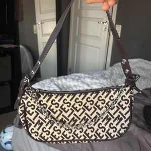 Beige och brun axelväska  - Säljer en snygg axelväska  i beige och brunt med tydligt logomönster. Väskan har justerbart axelband i brunt skinn och en silverfärgad kedjedetalj framtill. Perfekt storlek för det viktigaste och stängs med dragkedja.