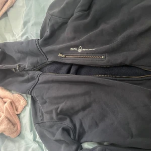 Svart Sail Racing fleecefodrad jacka M - Svart jacka från Sail Racing i storlek M med fleecefoder och dragkedja framtill. Jackan har en extra dragkedjeficka på bröstet och ribbade muddar vid ärmslut. Perfekt för kyliga dagar och har en sportig vibe. Lite sönder vid armarna där ni kan se på bilden , köpt för 1300kr.