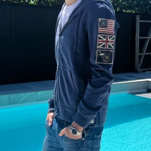 Ralph Lauren Denim & Supply - Snygg Ralph lauren Marinblå hoodie med dragkedja och huva, dekorerad med coola patchar på ena ärmen (USA, UK och flygpatch). Jackan har fickor framtill och snörning i huvan. Perfekt för en avslappnad streetstil. Priset kan diskuteras vid snabb affär