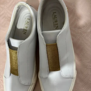 Vita sneakers från Lauren Ralph Lauren - Stilrena vita sneakers i skinn från Lauren Ralph Lauren med bred guldfärgad resårdetalj framtill och guldfärgad RL-logga på hälen. Skorna har platt sula och rund tå, perfekt för en clean och modern look.