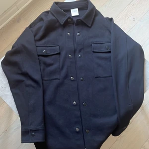 Svart overshirt från Selected Homme - Stilren svart overshirt från Selected Homme med klassisk krage, tryckknappar och två stora bröstfickor med lock. Jackan är i ett grovt bomullstyg och har långa ärmar med knappdetalj vid mudden. Perfekt för lager-på-lager och en clean look. Iprincip bara testad så 10/10 skick!