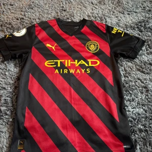 Manchester City Haaland matchtröja Puma - Snygg Manchester City bortatröja från Puma med Haaland och nummer 9 på ryggen. Svart bas med röda diagonala ränder och gula detaljer, inklusive klubbmärke och sponsortryck. Kortärmad modell i lätt och ventilerande material, perfekt för fotbollsfans.