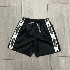 Svarta Puma shorts med logga - Svarta shorts från Puma med vita sidor och stor svart logga längs benen. Resår i midjan och snörning för perfekt passform. Lätt och luftig mesh på insidan, perfekta för sport eller chill. Klassisk streetstil med tydlig branding.