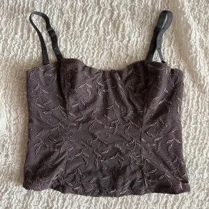 Mörkbrun broderad korsett-topp - Supersnygg mörkbrun korsett-topp med tunna, justerbara axelband. Framsidan har ett broderat blommönster i liknande ton som ger en lyxig känsla. Toppen är croppad och har en lätt figurnära passform. Perfekt att styla med jeans eller kjol för en trendig look.