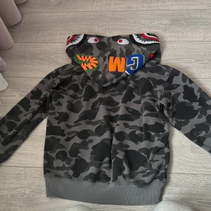 BAPE camo hoodie  - Säljer en grå och svart camo hoodie från BAPE med ikonisk shark-hood och broderade detaljer i färg. Tröjan har dragkedja hela vägen, huva och långa ärmar. Perfekt för dig som vill sticka ut med streetwear-vibe. 