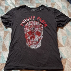Svart t-shirt med döskalle från Philipp Plein - Svart t-shirt från Philipp Plein med stort döskalletryck i rött och silver framtill. T-shirten har korta ärmar, rund halsringning och coola detaljer med logga på sidan och nacken. Materialet är mjuk bomull och passformen är lite mer som small skulle jag säga
