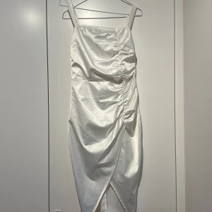 Vit satin klänning med drapering - Snygg vit långklänning i satin med omlott-detalj och drapering framtill. Klänningen är ärmlös med breda axelband och har en elegant glansig finish. Perfekt för dig som vill sticka ut med en stilren look.