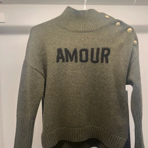 Olivgrön stickad tröja Zadig & Voltaire - Olivgrön stickad tröja från Zadig & Voltaire med hög krage och snygga guldknappar längs ena axeln. Svart broderad text 'AMOUR' framtill. Ribbade muddar och avslut, perfekt för lager-på-lager under kyliga dagar. Ganska  liten i storleken