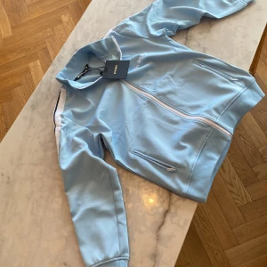 Ljusblå track jacket från Palm Angels - Säljer en ljusblå track jacket från Palm Angels med vit dragkedja och vita detaljer längs ärmarna. Jackan har två fickor med dragkedja och broderad logga på bröstet. Tillverkad i mjuk polyester för en sportig vibe.