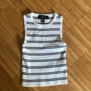 Randig ribbad linnetopp Forever 21 - Vit och svart ribbad linnetopp från Forever 21 med horisontella svarta ränder. Ärmlös och med rund halsringning, perfekt för en chill och stilren look. Toppens material är stretchigt och mjukt, vilket gör den superbekväm att bära.