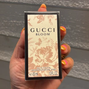 Gucci Bloom Eau de Toilette 30ml - Gucci Bloom Eau de Toilette i 30 ml flaska. Förpackningen är beige med blommigt och fjärilsmönster i ljusrosa toner och svarta kanter. En feminin och elegant parfym med stilren design, perfekt för dig som gillar lyxiga dofter.
