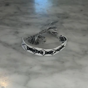 Märkesarmband svart - Snyggt Märkesarmband i grått och vitt tyg med svart broderad logga. Armbandet har ett grafiskt rutmönster och avslutas med flätade band och tofsar. Perfekt accessoar för att lyfta din outfit med en exklusiv touch.