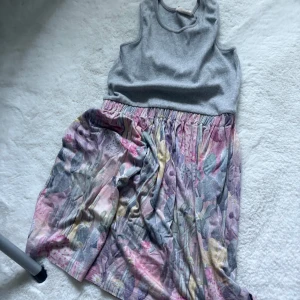Grå och blommig kortklänning - Supersöt kortklänning med grå ribbad överdel och blommigt mönstrad kjol i pastelliga toner av rosa, lila och gul. Ärmlös modell med rund halsringning och mjukt, bekvämt material. Perfekt för en somrig look!