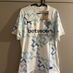 Inter Milan vit/blå Nike fotbollströja - Inter Milan fotbollströja från Nike i vitt med ljusblå och blå geometriskt mönster. Tröjan har klubbmärke och sponsortryck på bröstet samt klassisk rund hals. Tillverkad i lätt och ventilerande polyester, perfekt för match eller träning.