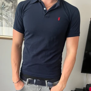 Marinblå pikétröja från Ralph Lauren - Snygg marinblå pikétröja från Ralph Lauren med klassisk krage och två knappar. Tröjan har korta ärmar och den ikoniska röda loggan broderad på bröstet. 9/10 skick, storlek S