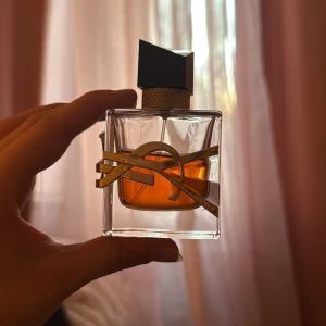 Yves Saint Laurent Libre Eau de Parfum - Snygg parfymflaska från Yves Saint Laurent med guldig metallogo runt flaskan. Flaskan är fyrkantig i genomskinligt glas med bärnstensfärgad vätska och har ett svart, asymmetriskt lock med gulddetaljer. Perfekt för dig som gillar lyxig design och ikoniska dofter.