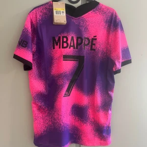 PSG Mbappé fotbollströja rosa/lila - Säljer en PSG fotbollströja med Mbappé 7 på ryggen. Tröjan har ett unikt rosa och lila mönster, svarta detaljer och klubbmärke på bröstet. Tillverkad i lätt och ventilerande material, perfekt för dig som vill sticka ut på planen eller läktaren.